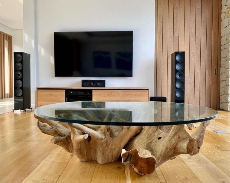 Modern living room featuring a large glass coffee table with a natural wood base, flanked by home theatre speakers, with a television mounted on the wall.