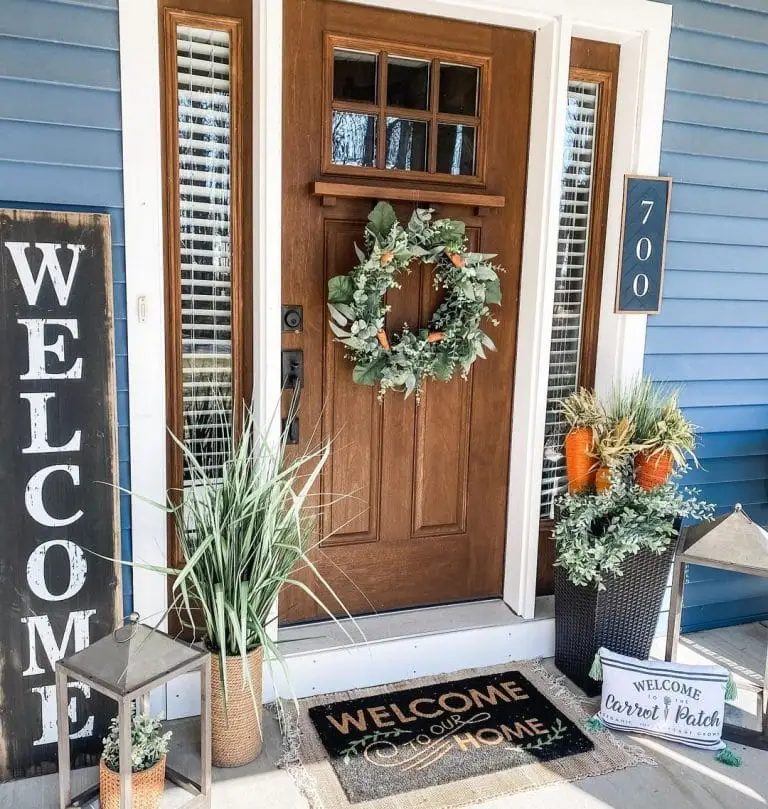 A natural wooden front porch door with a door wreath, a welcome sign, a welcome door mat, and potted flowers ready for the Spring season.