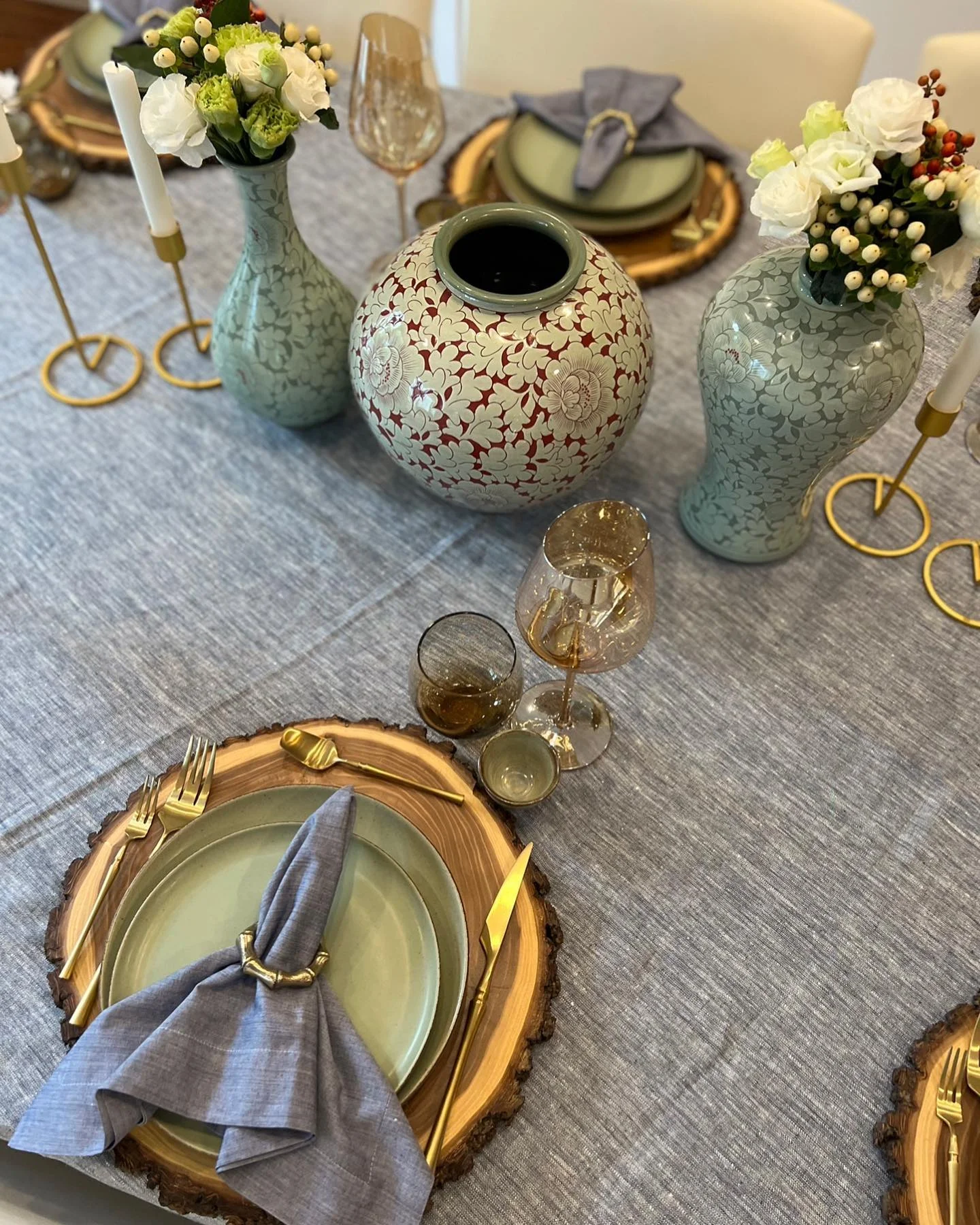 A table setting with floral vases, candles, layered plates, gold cutlery, glasses, and napkins on wooden chargers over a gray tablecloth exudes rustic charm with the addition of wood stumps as unique table decor.