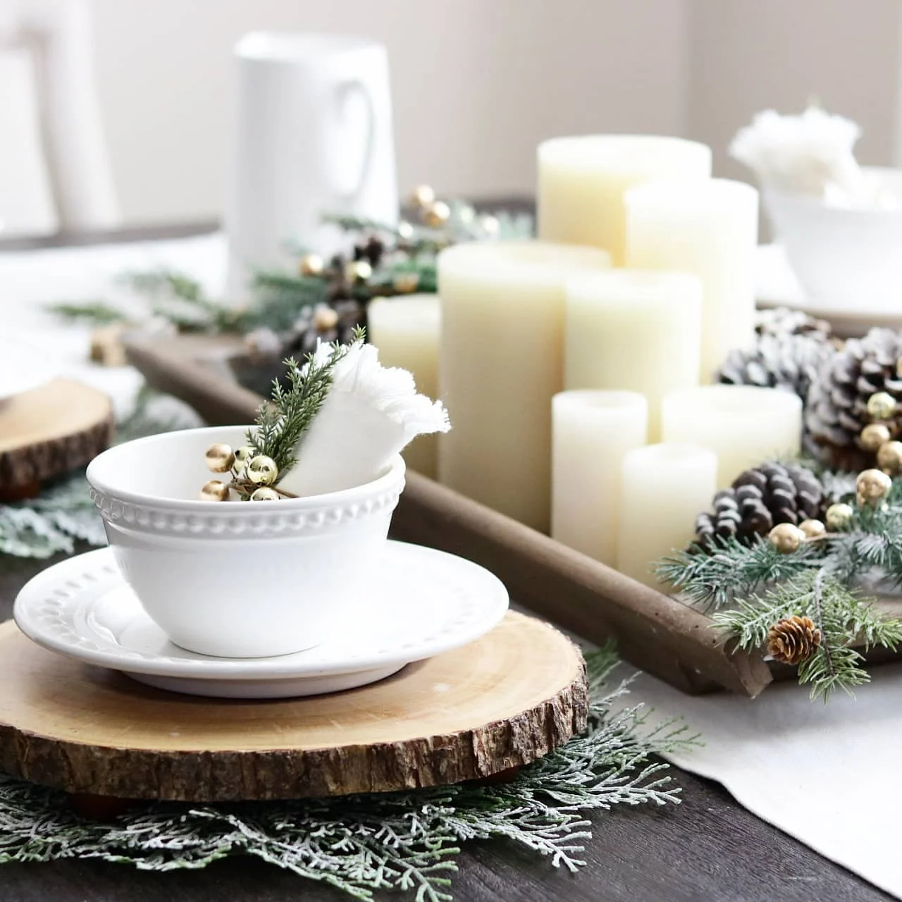 A white tea set resting on a wooden slice, surrounded by festive decor including candles, greenery, and pinecones on a dining table exudes rustic charm with its wood stumps table decor.