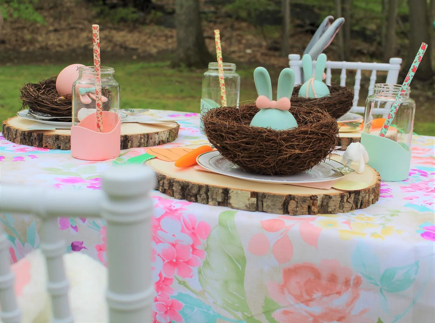 A table set for an outdoor gathering with floral tablecloths, mason jars with straws, and bird nests holding pastel-colored bunny-shaped eggs as centerpieces. White chairs surround the table, accented by rustic charm wood stumps as additional decor.