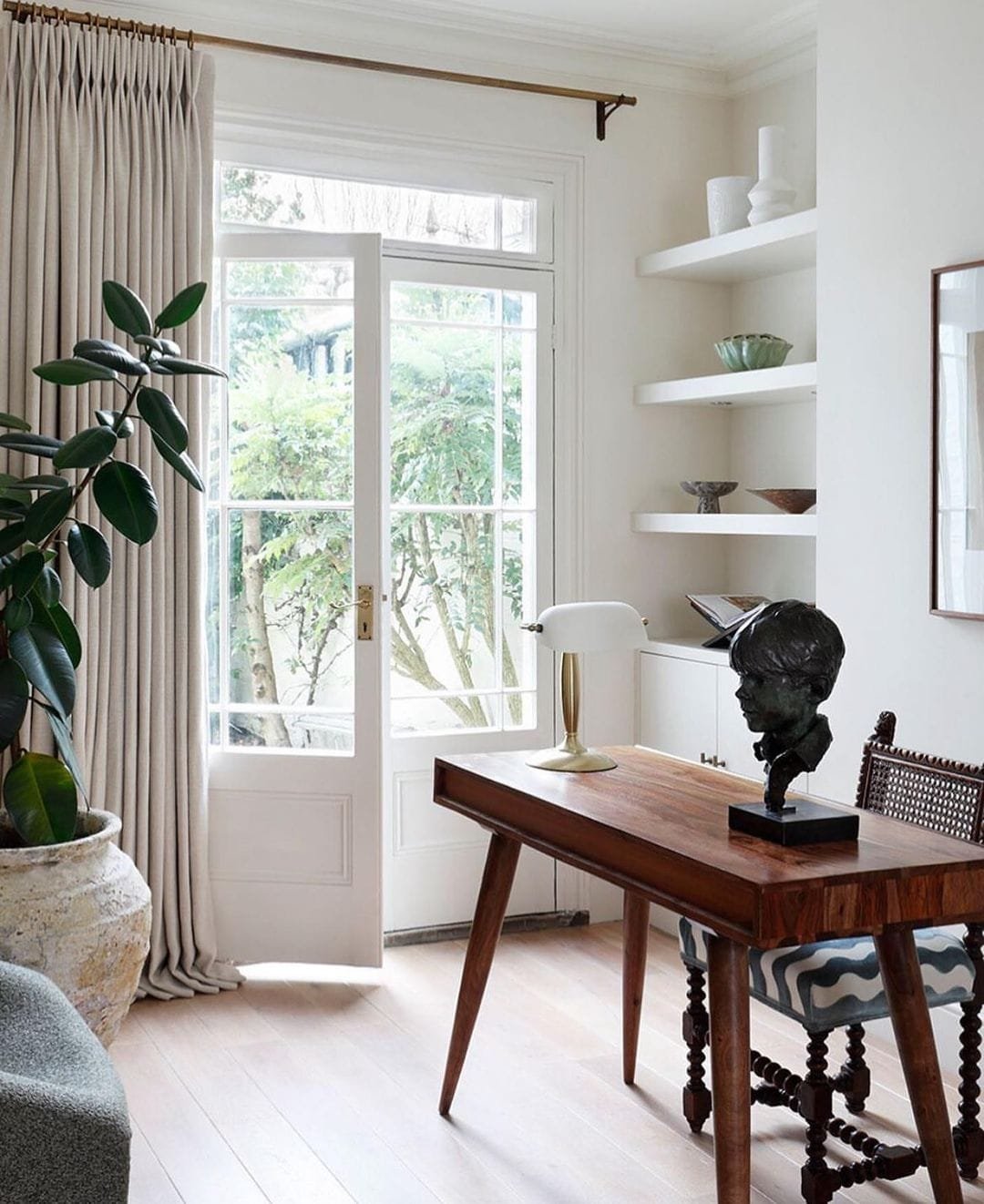 Bright room featuring a wooden desk, classic chair, and a small sculpture. A large plant and built-in shelves near a window with sheer curtains provide practical ways to add natural elements to your home.