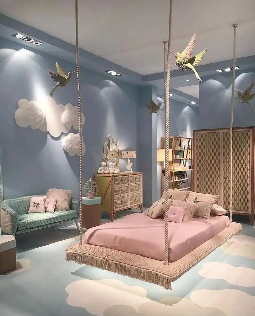A whimsical bedroom with a hanging bed framed by ropes, cloud decorations on blue walls, bird sculptures, a small couch, and a dresser filled with plush toys creates one of the 17 pink bedrooms fit for a princess.