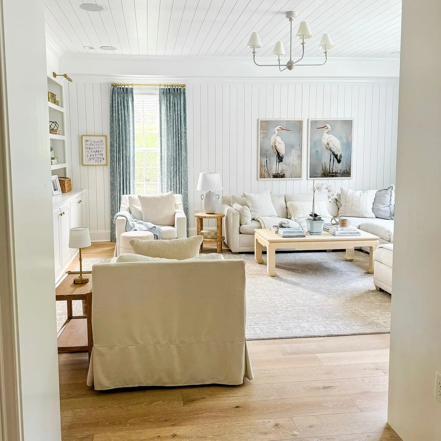 Bright living room with white walls, light wood floors, and neutral furniture features a large sectional sofa, coffee table, built-in shelves, and two bird prints above the couch—perfect for coastal-inspired decor to bring the beach to your living room.