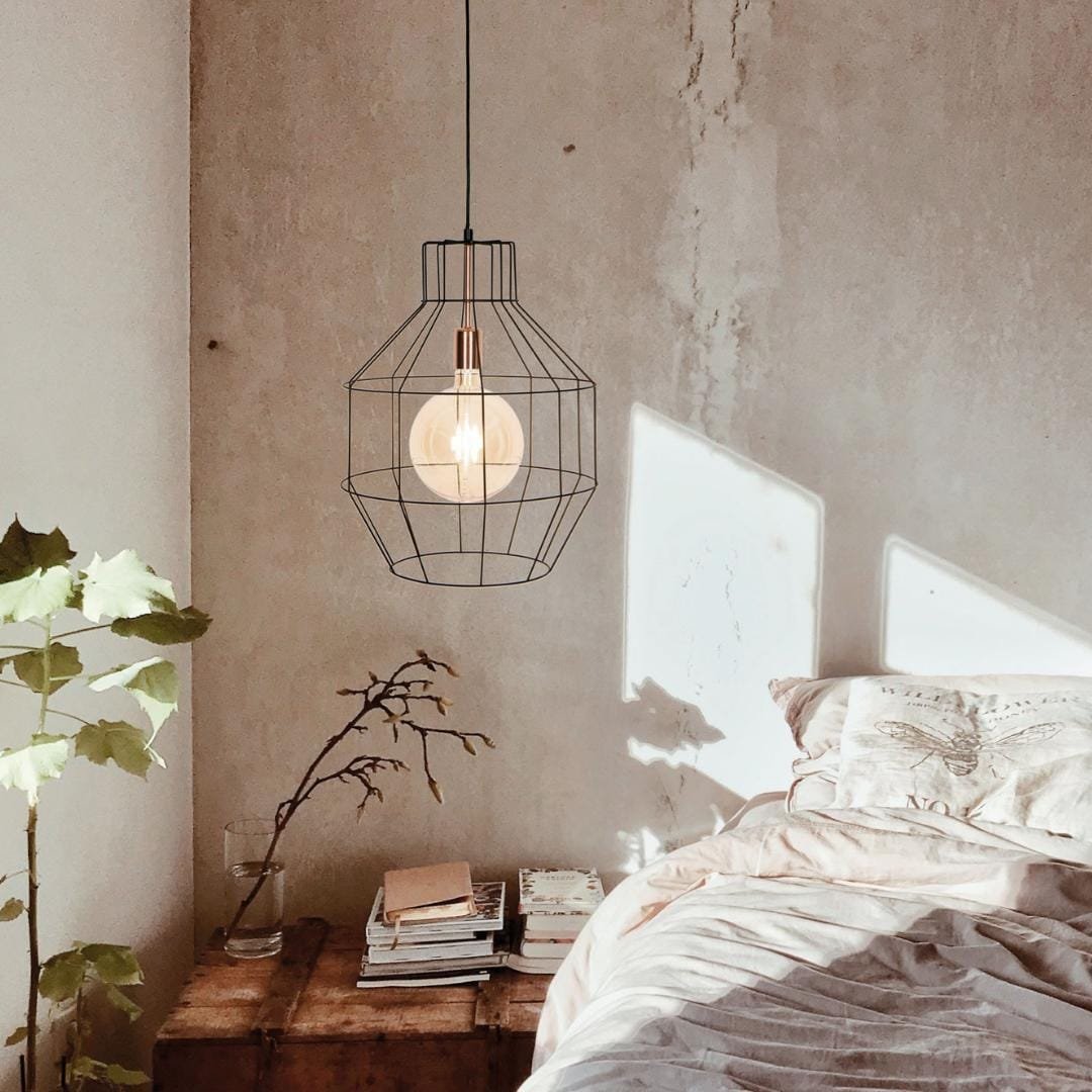 A cozy bedroom corner featuring an unmade bed, a hanging geometric lamp, and an industrial chic wooden bedside table with books and a small potted plant.
