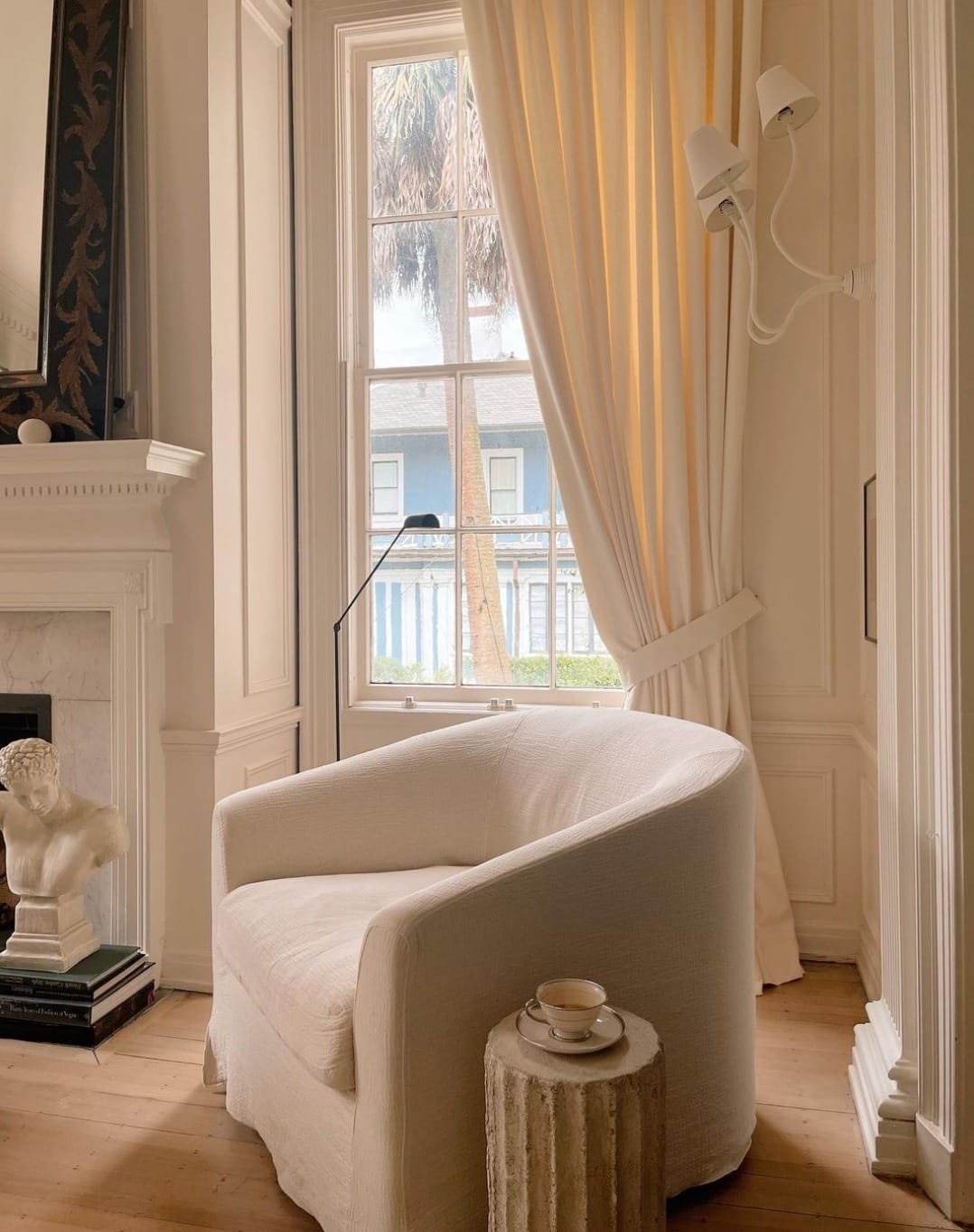 Elegant minimalist living room with a white sofa, decorative fireplace, and draped window showcasing a palm tree view.
