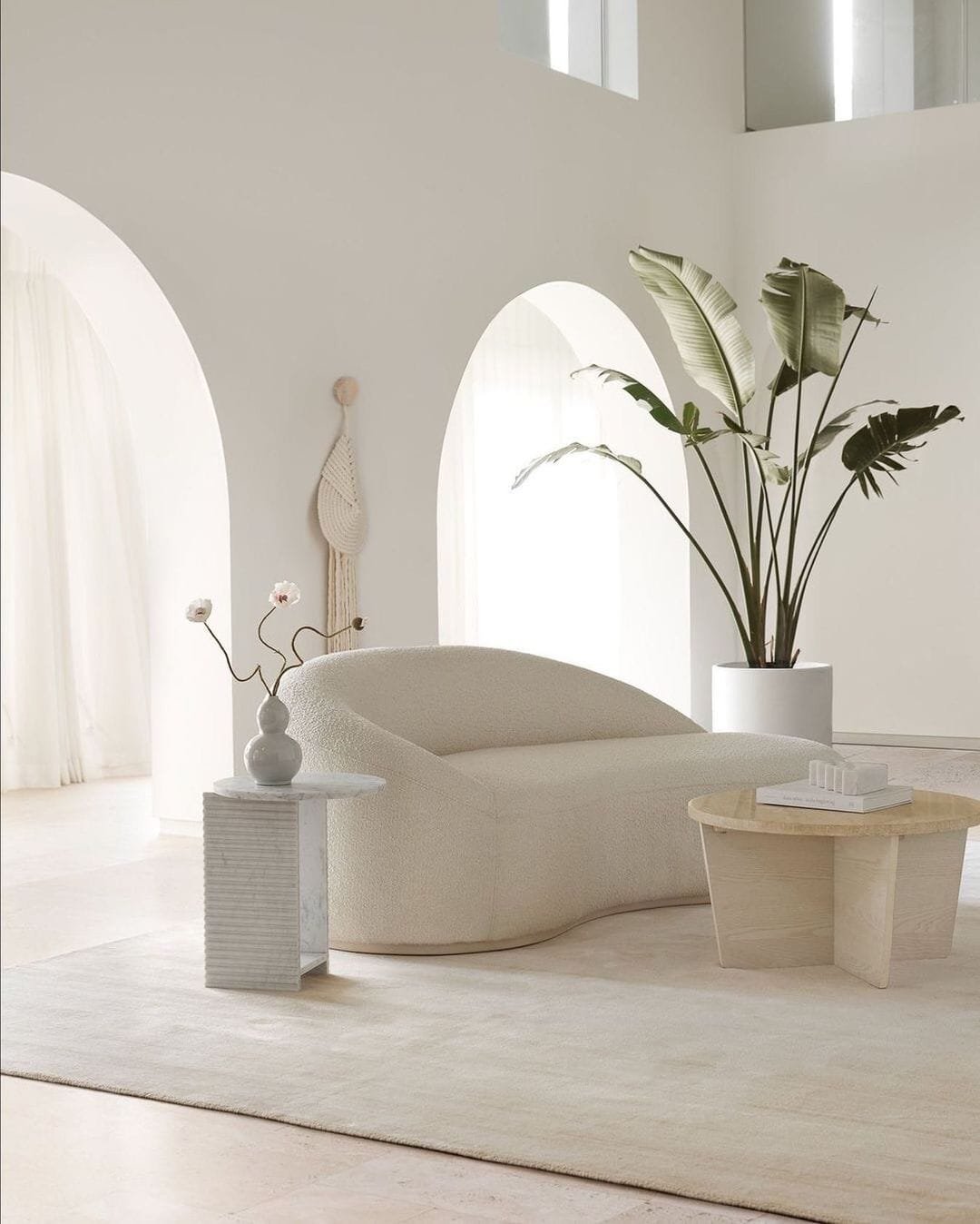 A minimalist living room with a plush white sofa, a small side table with decorative items, and a potted tropical plant, set against arched white walls and sheer curtains. Discover our top 10 tips for creating this serene space.