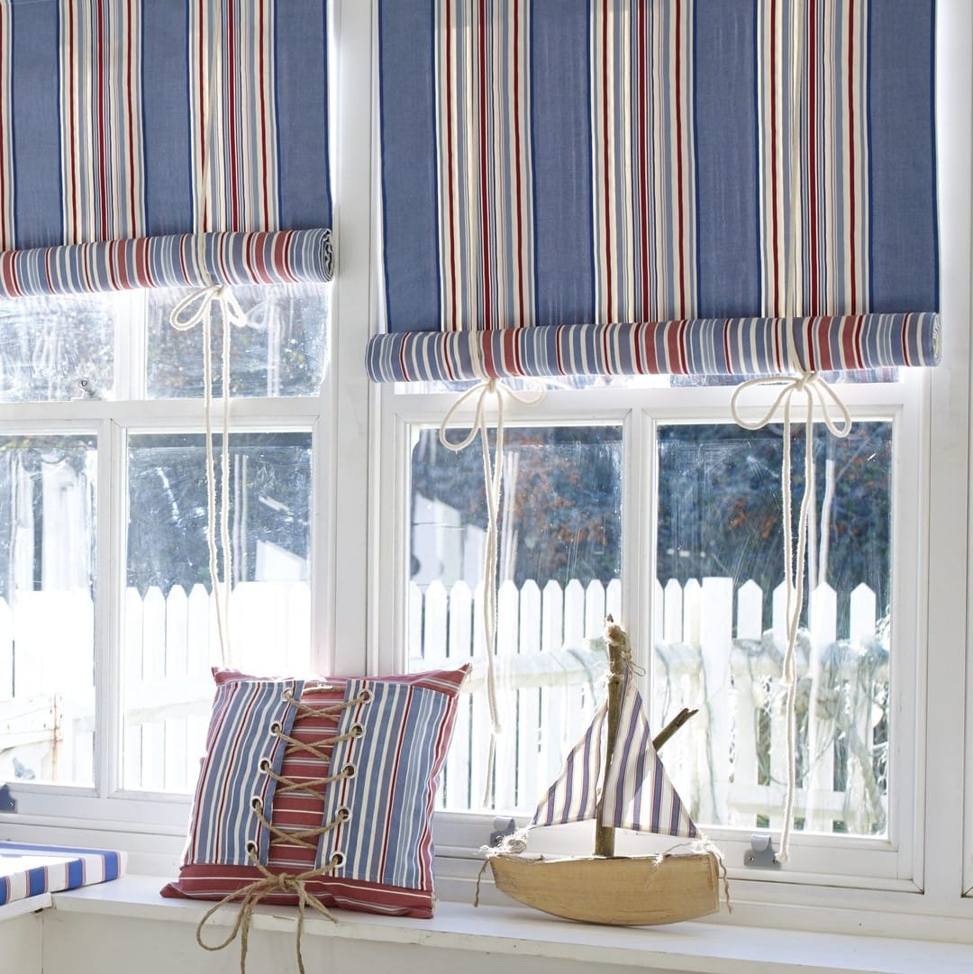 Coastal-inspired window with striped Roman shades and cushions, toy sailboat, in a sunny, white-framed window.