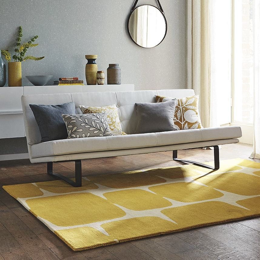 A modern living room featuring a white sofa with funky cushions, a yellow and white rug, and a round mirror on a textured wall.