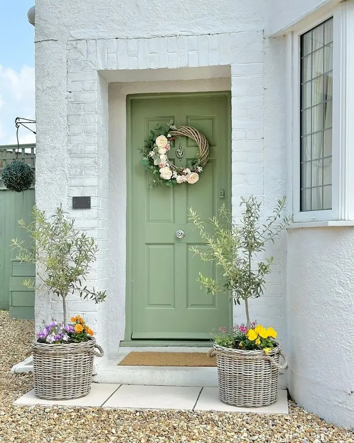 A pale green front door with a floral wreath, flanked by two wicker baskets—explore the charm of wicker and discover Rattan vs. Wicker: Which is Better for Your Summer Decor?—all set in a white brick house with colorful blooms.