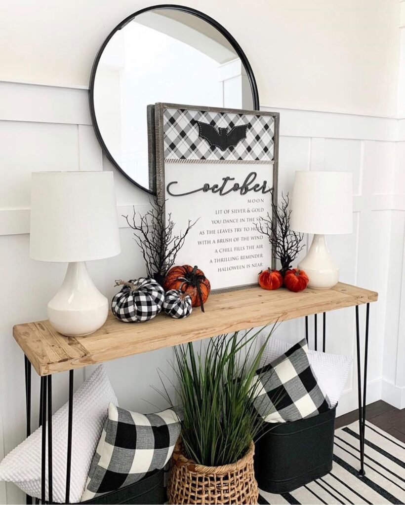 A wooden console table with white lamps, autumn decor, plaid pumpkins, and a sign reading "October" sits below a round mirror. Baskets and pillows underneath show how to embrace buffalo plaid in fall decor for a cozy seasonal vibe.