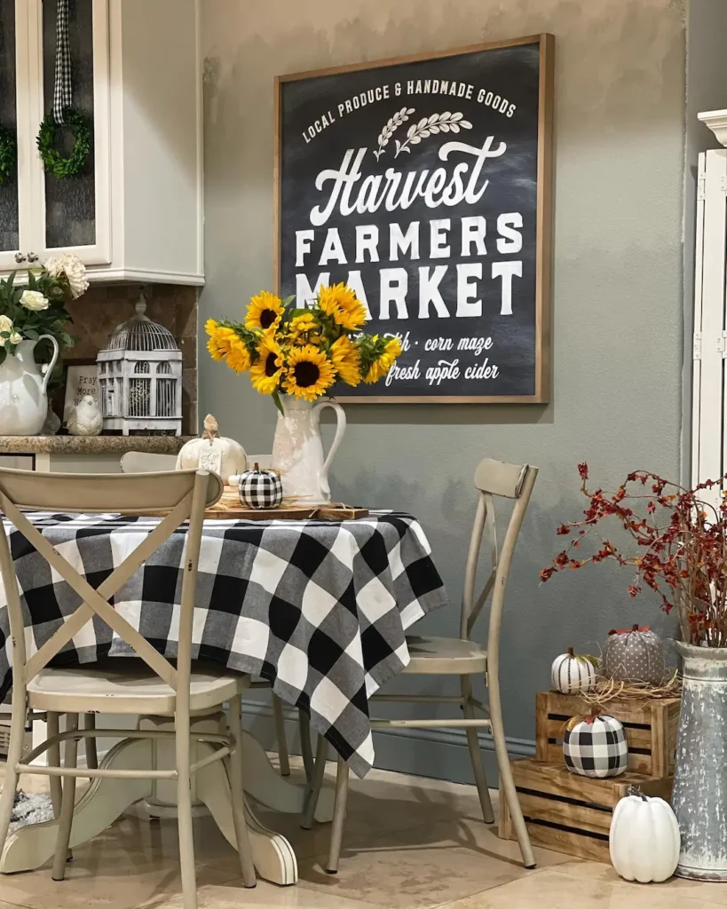 A small dining area with a checkered tablecloth—showing how to embrace buffalo plaid in fall decor—sunflowers in a pitcher, decorative pumpkins, and a "Harvest Farmers Market" sign on the wall.