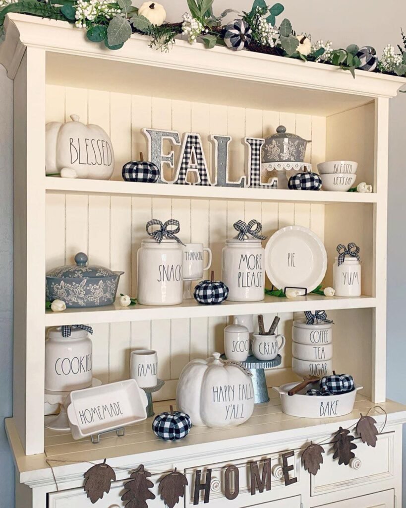 A white hutch decorated with fall-themed ornaments, ceramic dishes labeled “FALL,” “BLESSED,” and “SNACKS,” plaid pumpkins, and garlands of leaves and greenery shows how to embrace buffalo plaid in fall decor for a cozy seasonal touch.