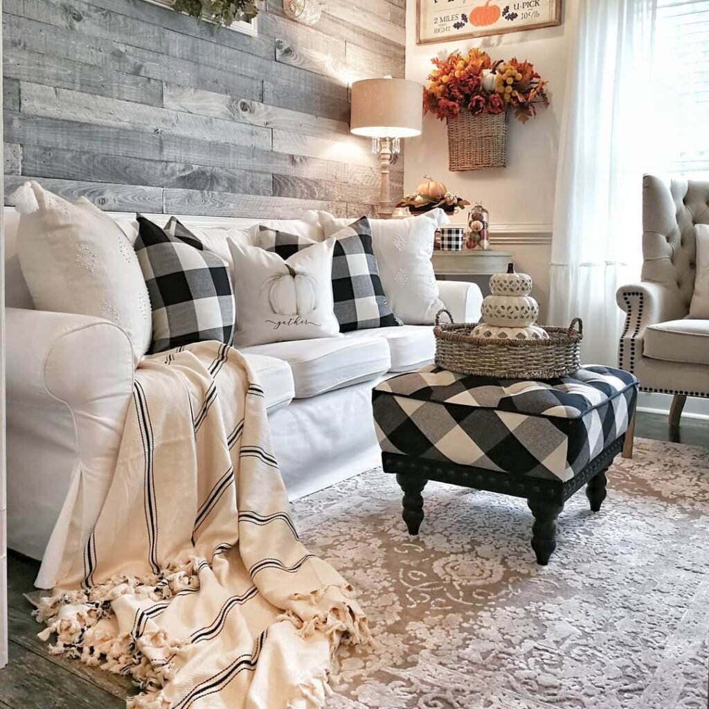 A cozy living room with a white sofa, plaid pillows, a beige throw blanket, and a plaid ottoman shows how to embrace buffalo plaid in fall decor with seasonal touches like a pumpkin sign and a basket of flowers.