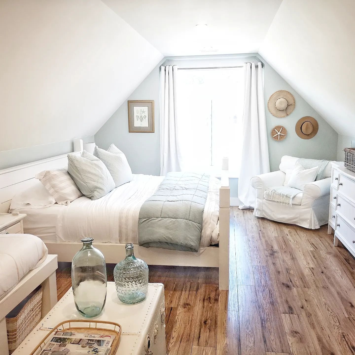 A bright, coastal attic bedroom with a large bed, white linens, two chairs, a small table with glass jars, and wooden floors, leading to a balcony with curtains.