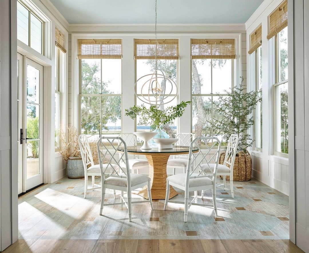 Bright dining room with large windows, a round wooden table, white chairs, a minimalist chandelier, and green plants.