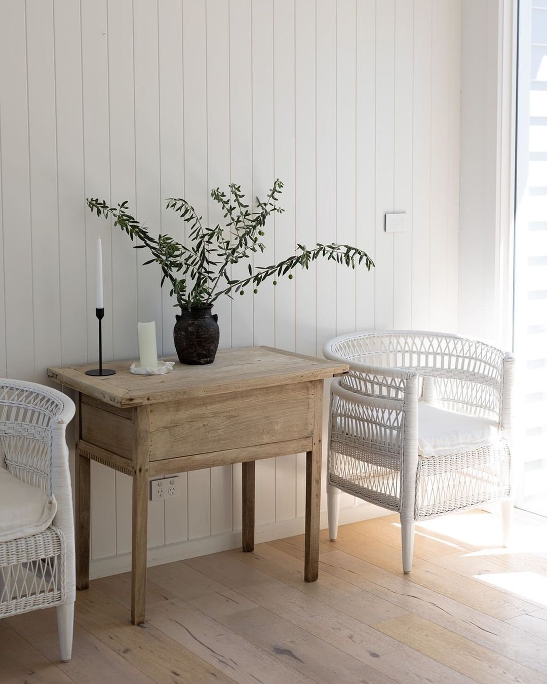 A minimalist dining area featuring a rustic wooden table, two white wicker chairs, and a vase with greenery, against a white paneled wall in coastal farmhouse decor.