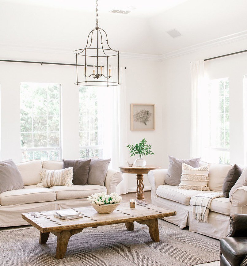 A cozy living room with a coastal farmhouse decor, featuring a large sectional sofa, a rustic wooden coffee table, and a modern hanging chandelier. White walls and bright windows enhance the airy vibe.
