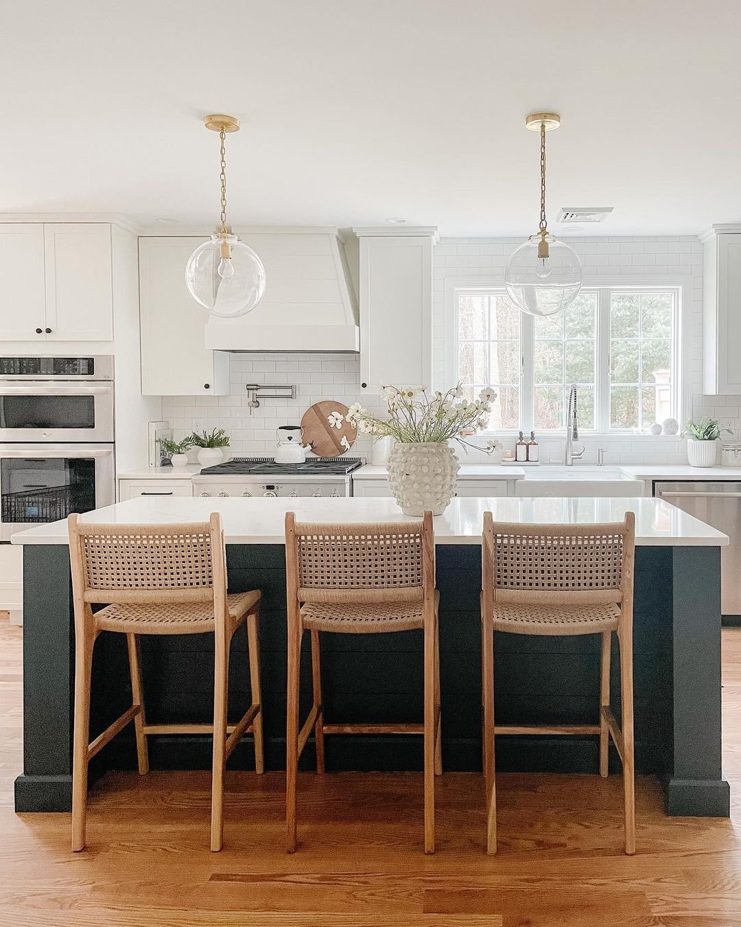 A modern kitchen with white cabinets, a navy blue island with rattan stools, gold pendant lights, and a vase of flowers showcases elements of Coastal Farmhouse decor.