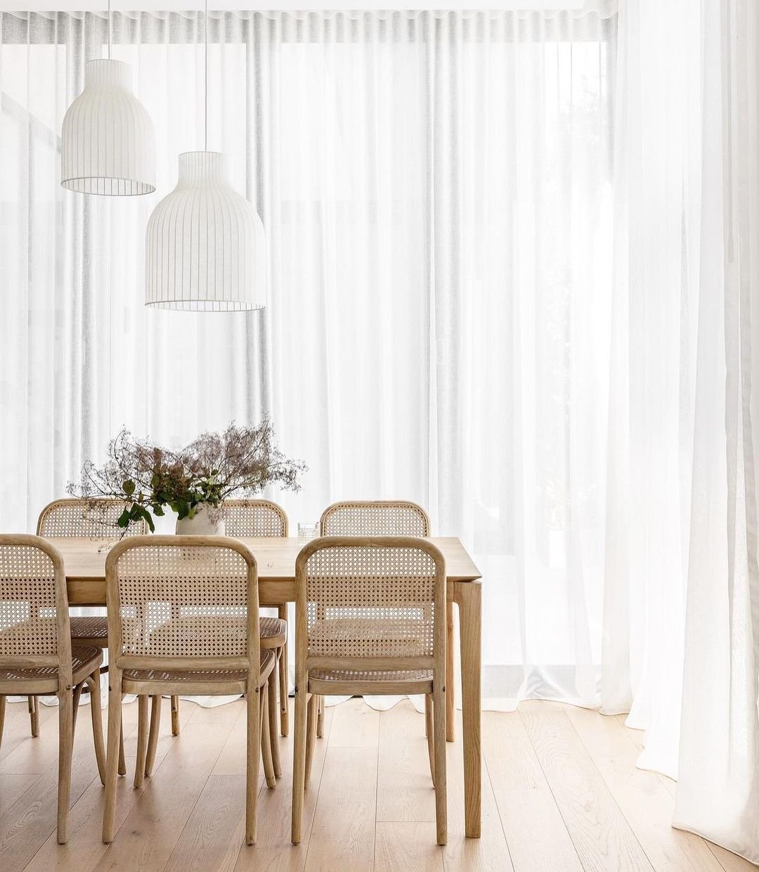 A bright, coastal dining room featuring a wooden table with matching chairs, white pendant lights, and sheer curtains.