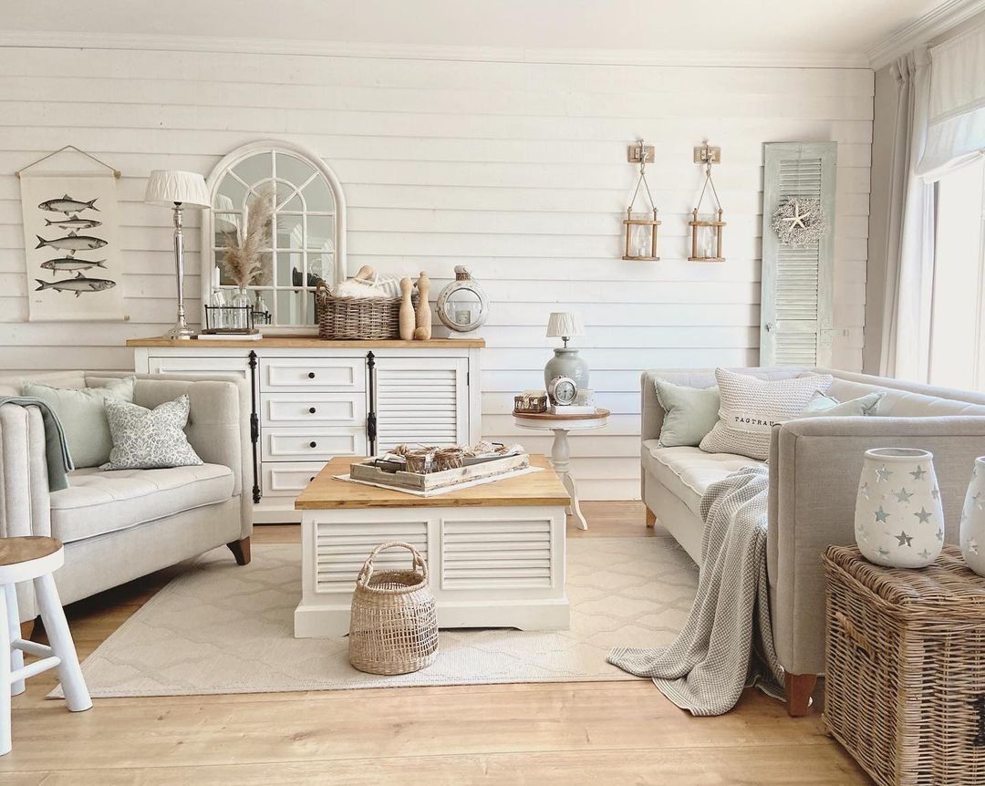 Cozy coastal-style living room with neutral tones, plush sofas, a minimalist wooden coffee table, woven baskets, and nautical decorations.