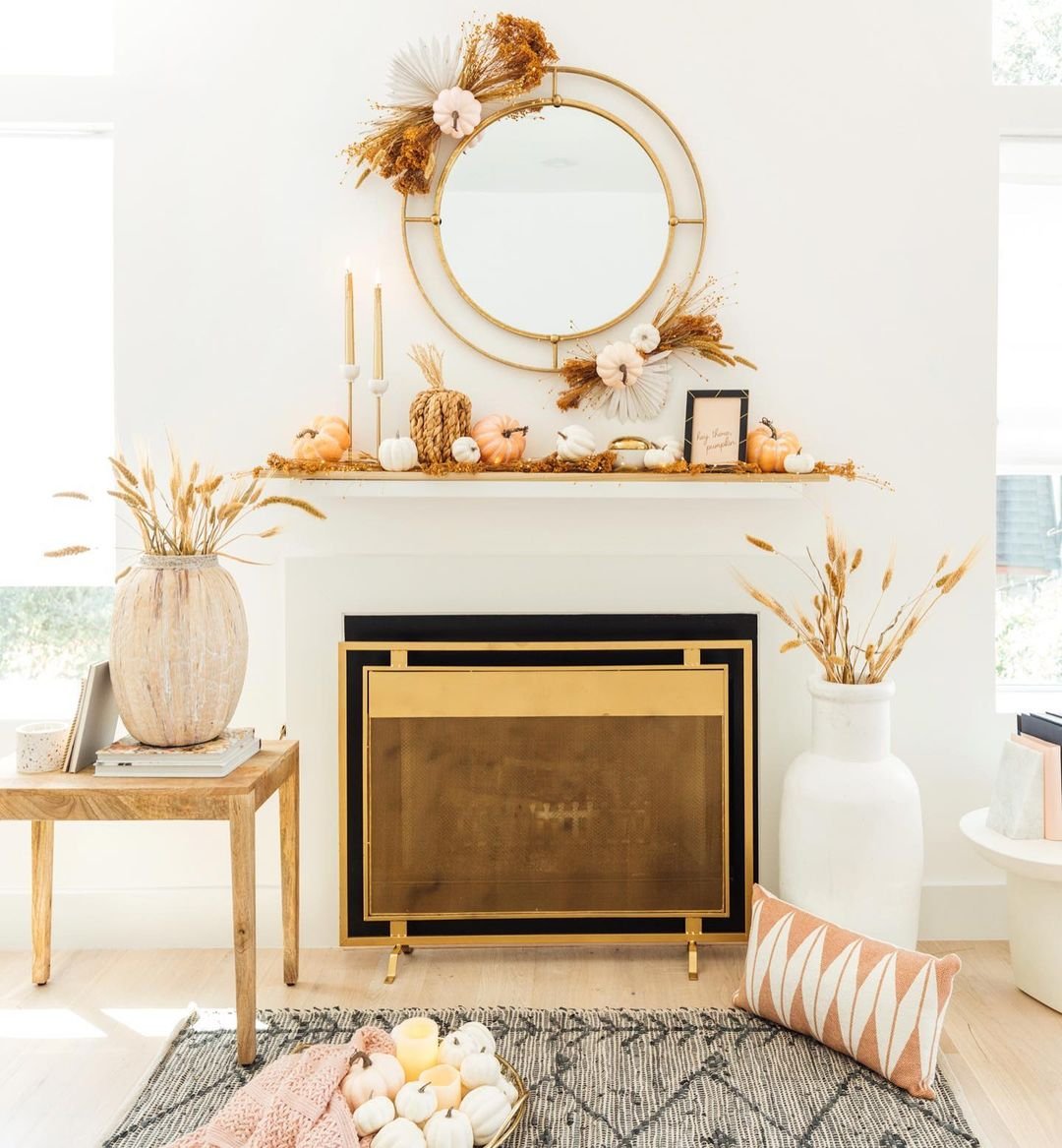A decorated fireplace with a round mirror, candlesticks, pumpkins, and wheat stalks on the mantel showcases neutral fall mantel decor ideas for a chic and sophisticated look. Nearby, a wooden table holds vases filled with wheat. A rug and throw pillows lie in front of the fireplace.