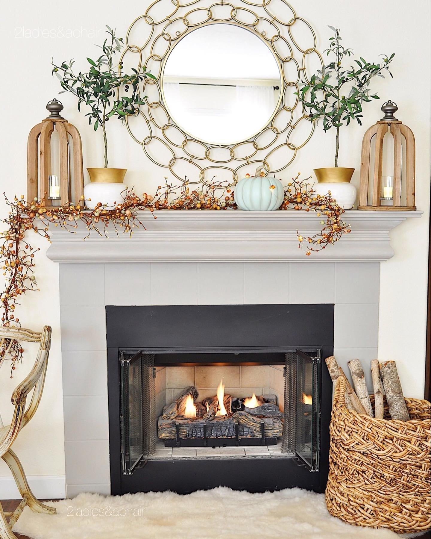 A decorated fireplace mantle with a round mirror, plants, a white pumpkin, and lanterns embodies neutral fall mantel decor ideas for a chic and sophisticated look. The fire is lit, casting a cozy glow, while a basket of logs rests elegantly beside the hearth.