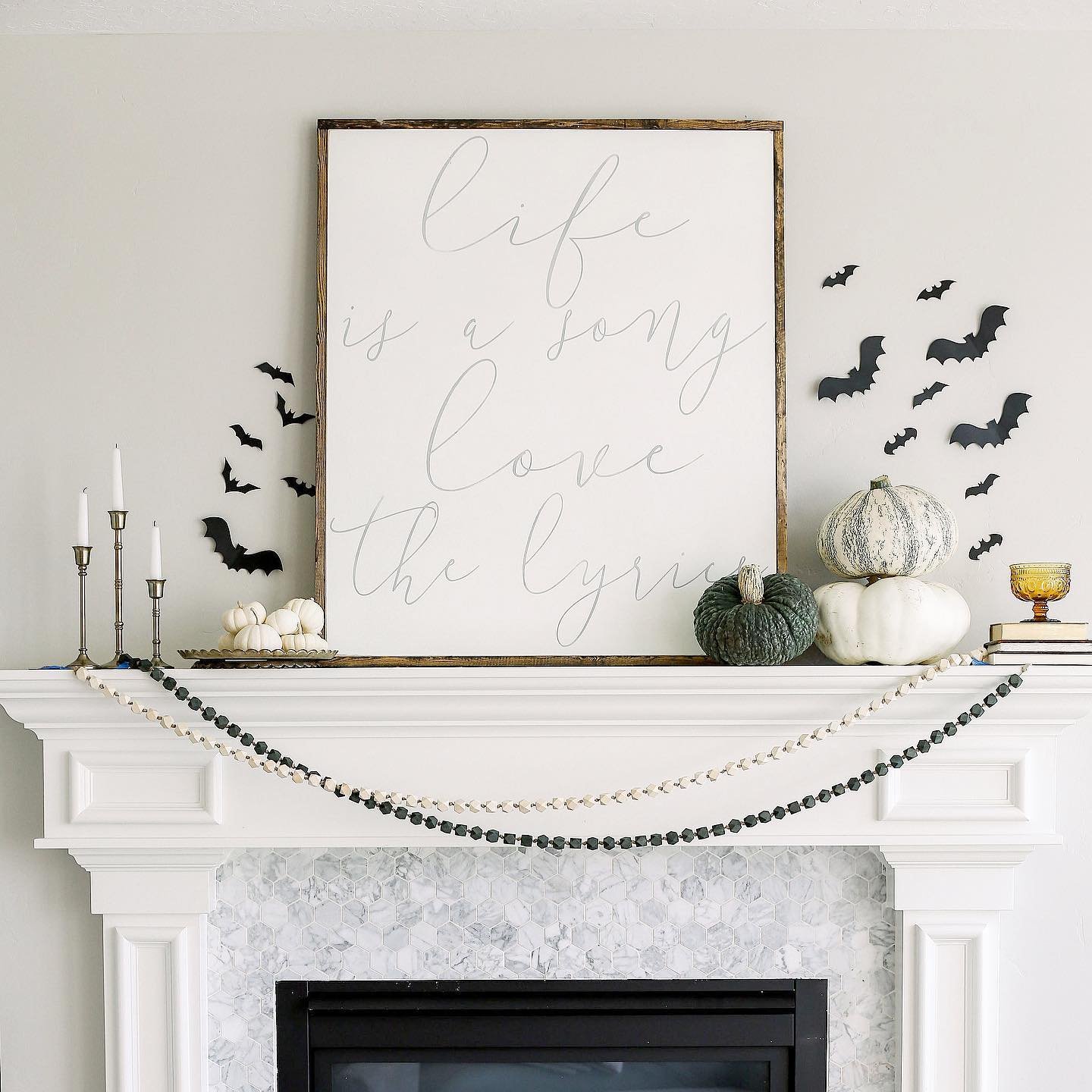 A white fireplace mantle decorated with pumpkins, candlesticks, a banner, and black bat cutouts offers neutral fall mantel decor ideas for a chic and sophisticated look. A framed sign above the mantle reads, "Life is a song, love the lyrics.
