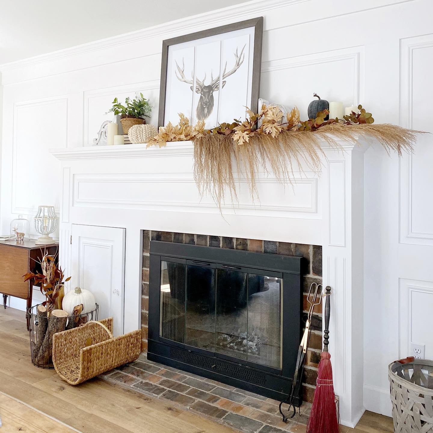 A white mantel decorated with autumn-themed foliage, a framed deer print, pumpkins, and candles offers neutral fall mantel decor ideas for a chic and sophisticated look. Below is a brick fireplace with accessories including a broom and fireplace tools on the right.