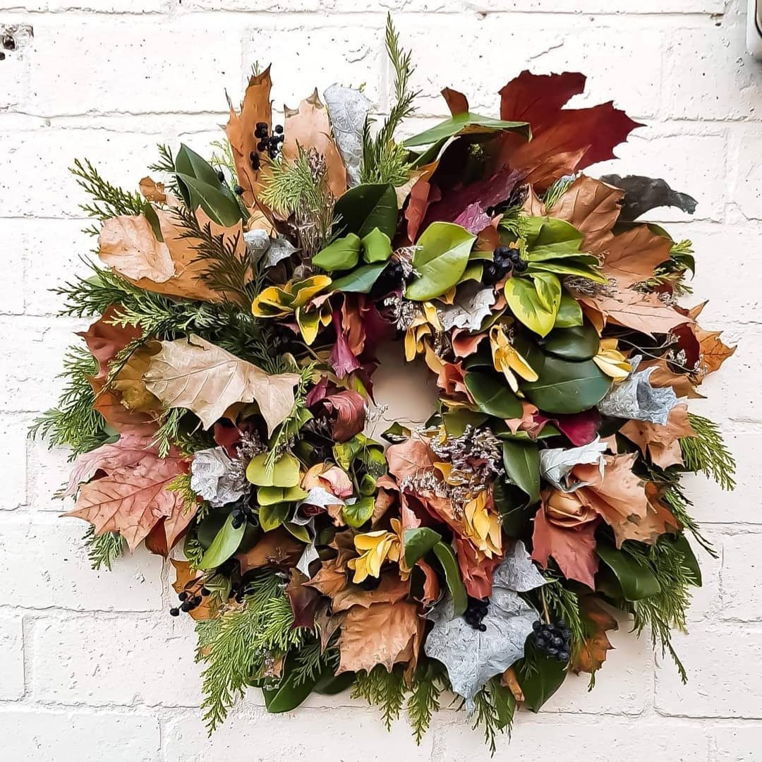 A decorative wreath made of autumn leaves and greenery is hung on a white brick wall, showcasing how to make your own fall wreaths with natural materials and scents.