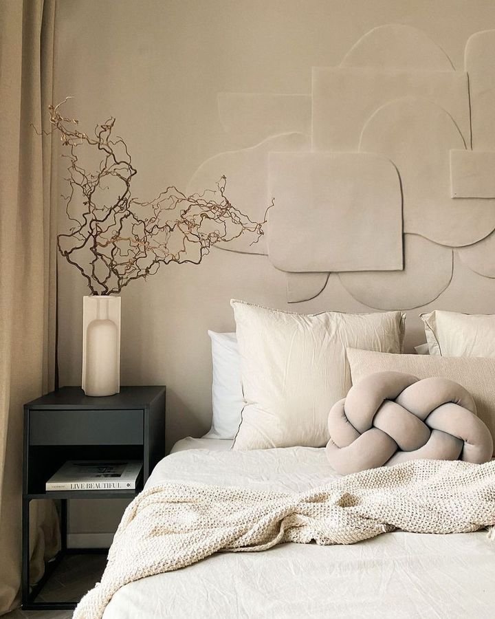 A minimalistic bedroom features a bed with neutral pillows and a knot cushion, embracing the principles of Minimalist Living: How Japandi Decor Can Help Simplify Your Life. A black nightstand with books and a vase holding branch decor complements the textured, abstract wall art in the background.