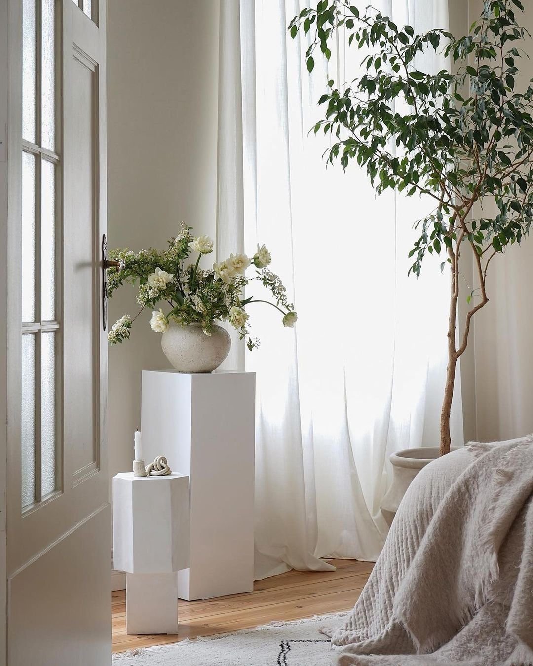 A minimalist bedroom embodies the essence of Japandi decor with a white vase holding flowers on a pedestal, a tall indoor plant, and sheer curtains allowing natural light to stream in. A bed with a beige blanket is partially visible, perfect for those embracing minimalist living.