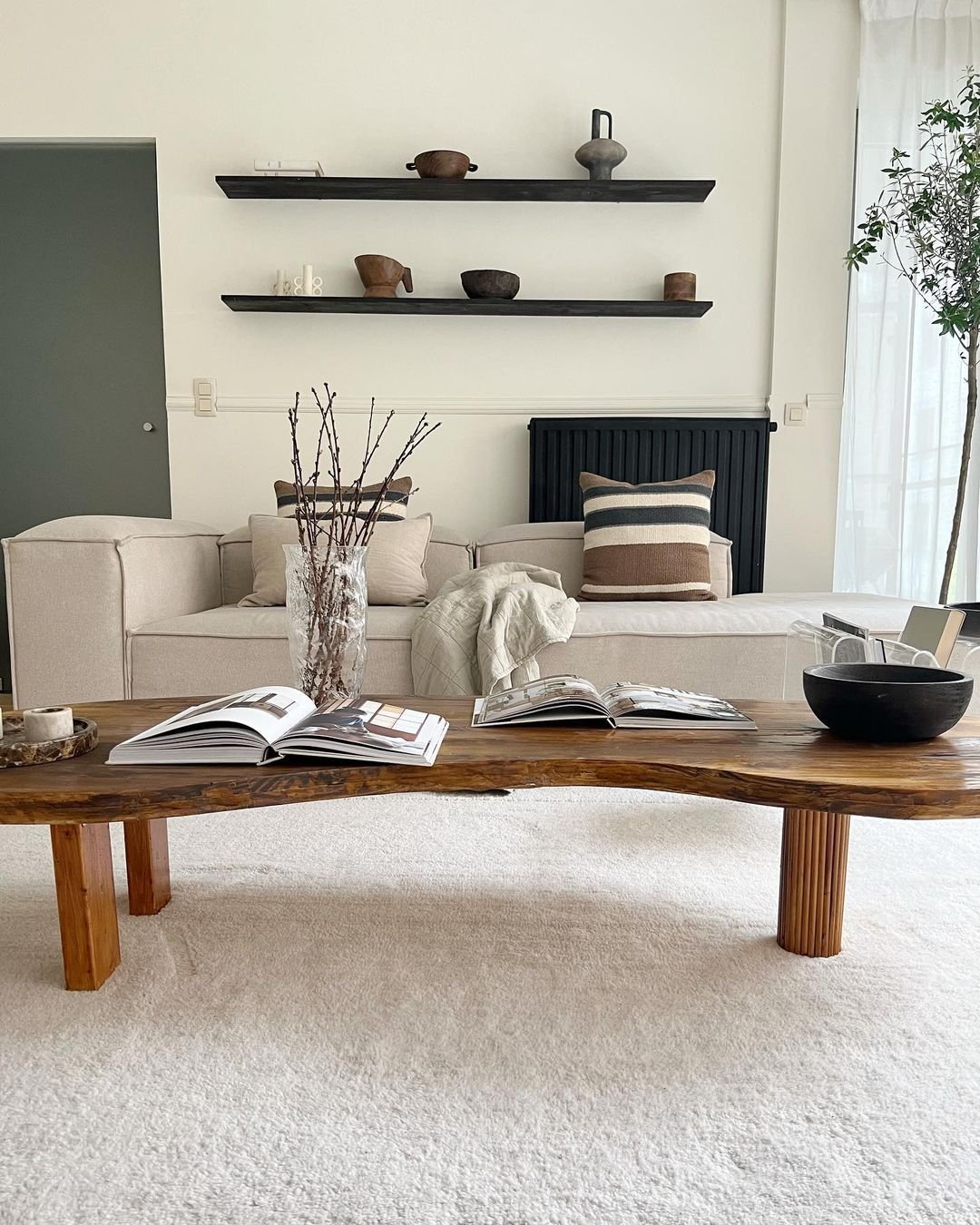 A modern living room with a white sofa, wooden coffee table with open books, and wall-mounted shelves holding pottery. Neutral colors dominate the decor, embracing the minimalist living that Japandi decor is known for, helping to simplify your life.