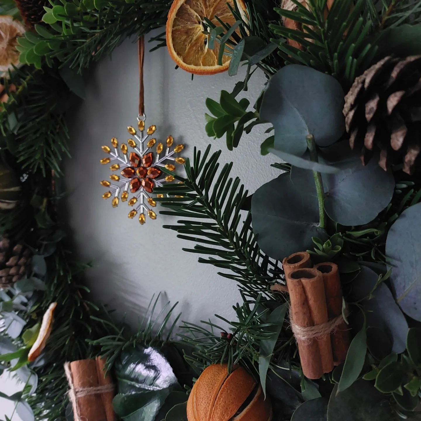A Christmas wreath decorated with dried orange slices, a beaded snowflake ornament, cinnamon sticks, pine cones, and green foliage hangs on a light-colored wall. Learn how to make your own fall wreaths with natural materials and scents for a touch of seasonal charm.