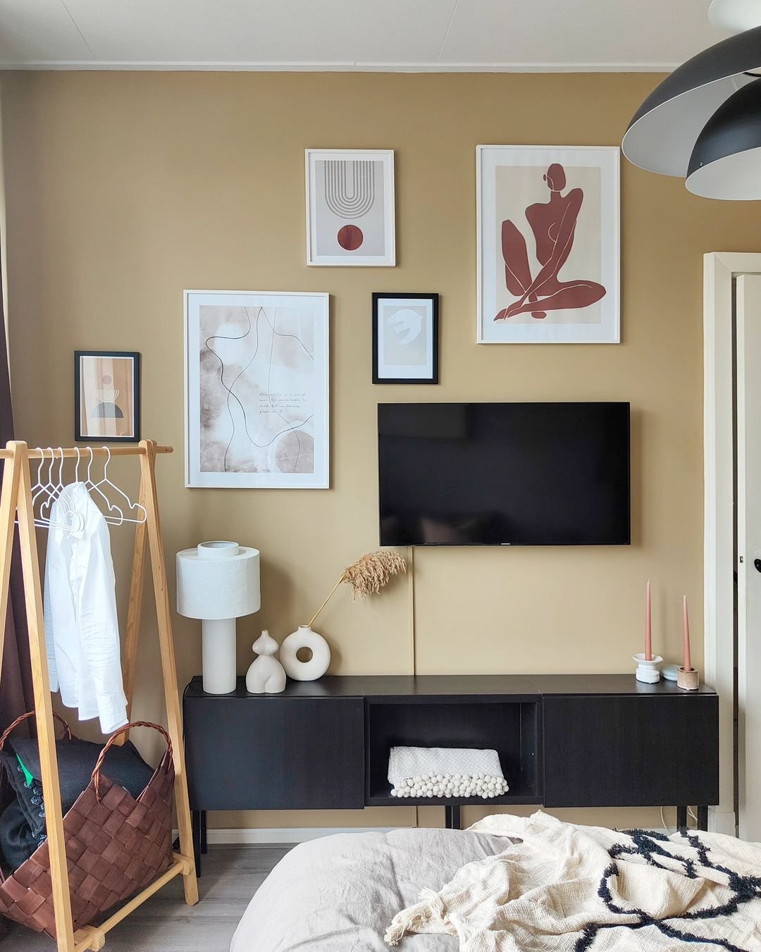A modern bedroom with a flat-screen TV mounted on a beige wall, various framed artworks, a black console table, a wooden clothing rack, and a bed with beige bedding. Discover how to incorporate meaningful artifacts in your Japandi bedroom for an elegant and personalized touch.