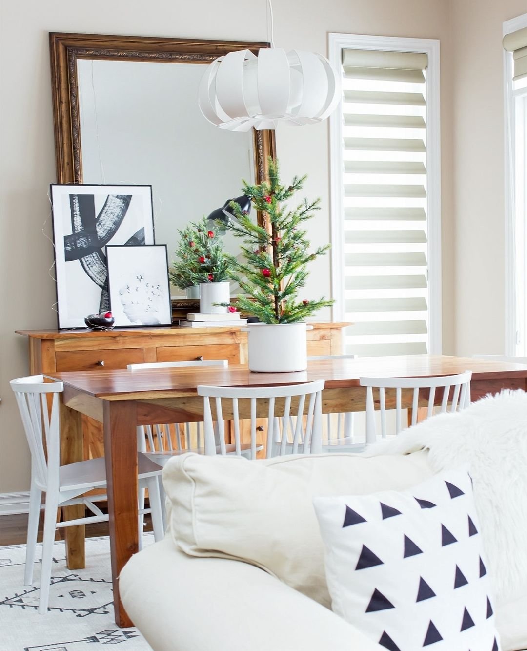 A modern dining area embracing minimalism features a wooden table, white chairs, and chic winter decor. A small potted Christmas tree sits on the table, while a large mirror and framed artwork adorn the wooden console against the wall.