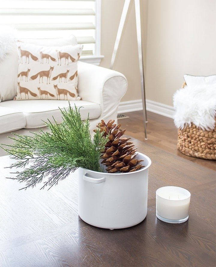 A white sofa with a patterned pillow sits in the background, next to a plant and large pine cone arrangement in a white container on a wooden table. Embracing minimalism, a chic winter decor scheme is completed with a lit white candle on the table.