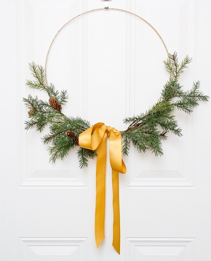 Minimalist wreath with pine branches, pinecones, and a yellow ribbon hangs on a white door, embracing minimalism for a chic winter decor scheme.