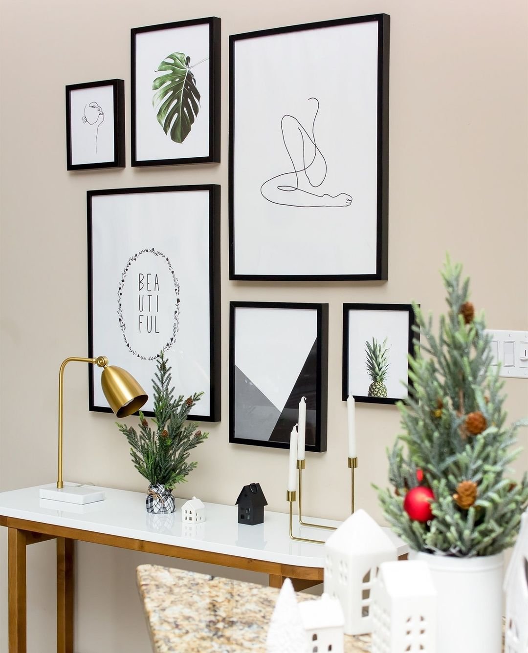 A collection of framed minimalist art, including abstract designs and plant illustrations, adorns a beige wall above a white console table. Embracing minimalism, the table also displays small decorative trees and candles, perfect for creating a chic winter decor scheme.