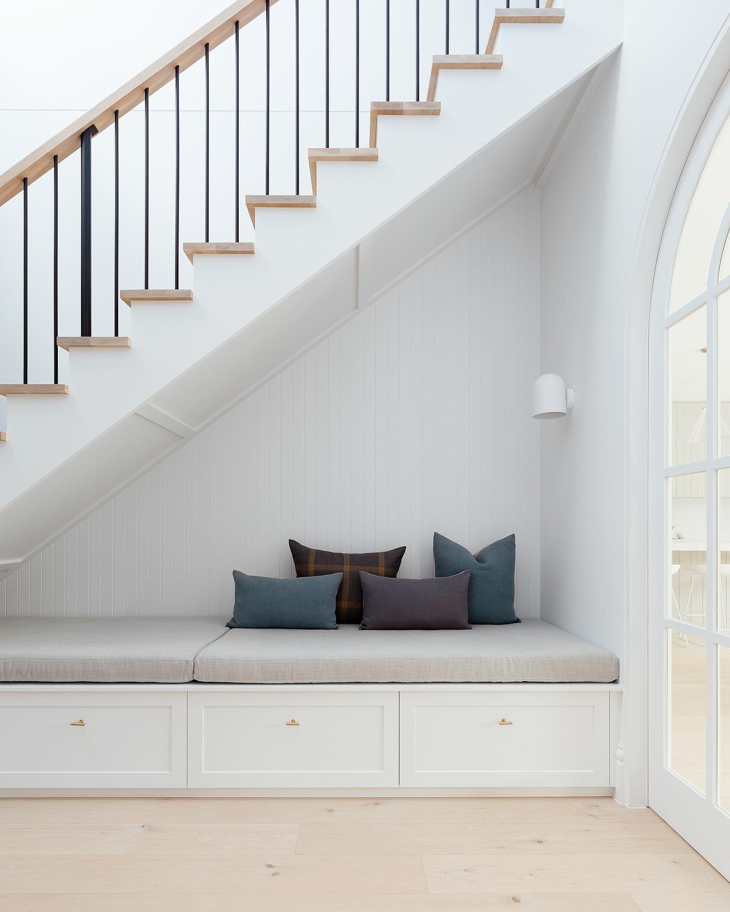 A small nook with a cushioned bench and pillows is tucked under a staircase. The space has white walls, a wooden floor, and a black metal stair railing. An arched doorway is visible on the right, showcasing one of the 10 Creative Under Stairs Storage Ideas to Optimize Your Space.