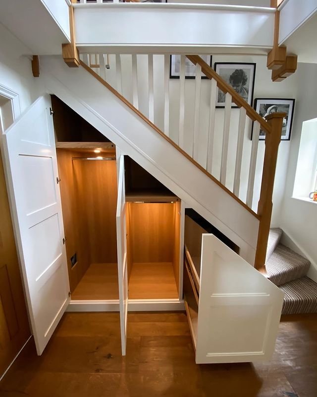 A view of an under-stair storage space with white panel doors open, revealing lighted wooden compartments. Stairs with white balusters and a wooden handrail are visible above in a well-lit area, showcasing the perfect under-stairs pantry: a dream storage solution for small kitchens.
