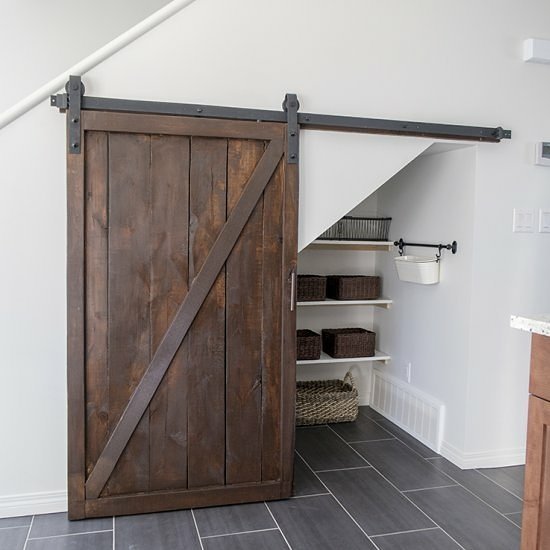A wooden sliding barn door opens to reveal an under-stairs pantry, a dream storage solution for small kitchens. Equipped with shelves holding baskets and storage containers, the cozy space maximizes your kitchen's efficiency. The floor is tiled in gray.