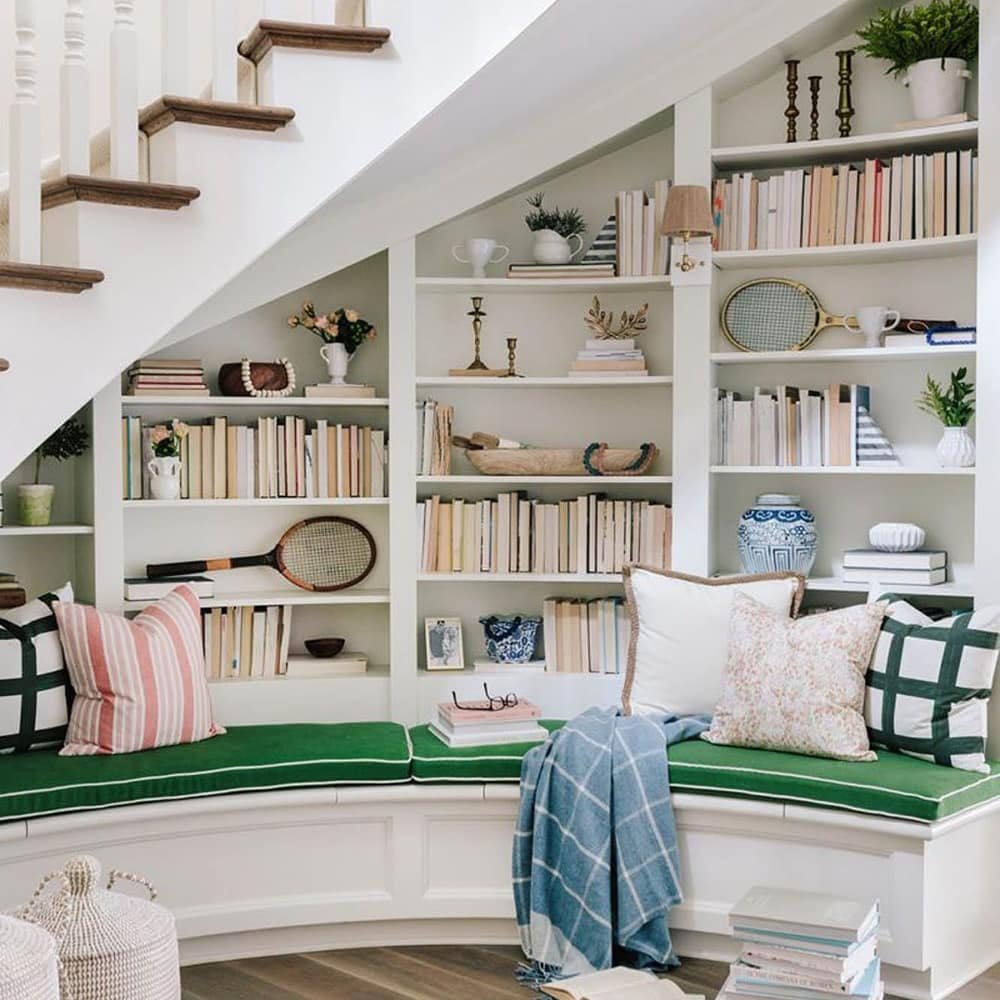 A cozy reading nook under a staircase features a built-in bookshelf filled with books and decorative items. The curved seating area is adorned with green and patterned cushions and a blue blanket, exemplifying one of the 10 Creative Under Stairs Storage Ideas to Optimize Your Space.