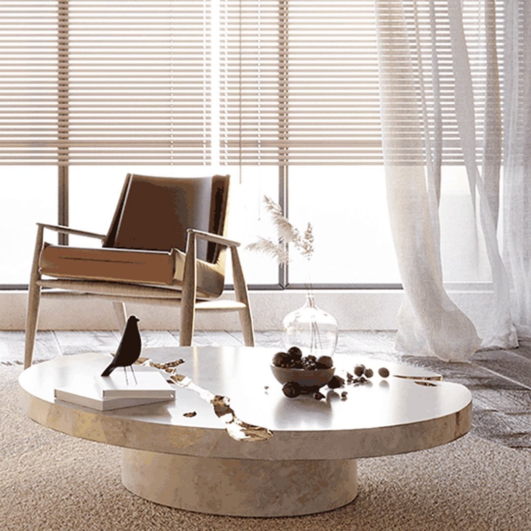 A modern living room features a circular coffee table with decorative items, a beige armchair, floor-to-ceiling windows with blinds, and sheer curtains that reflect the beauty of imperfection, resembling the Wabi-Sabi philosophy often seen in Japandi bedroom decor.