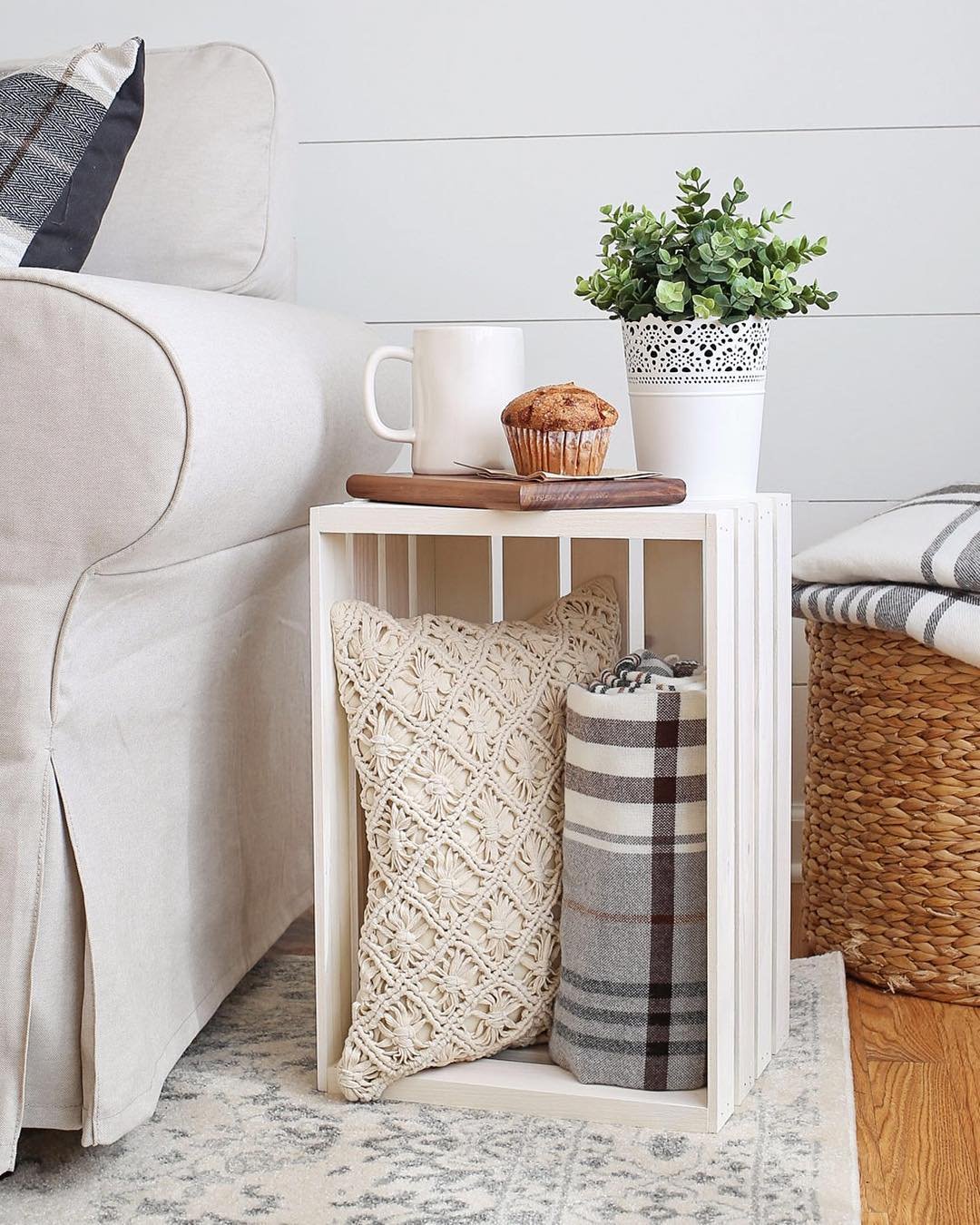 A wooden crate by a sofa holds a blanket and a crocheted pillow. It's topped with a tray holding a mug, muffin, and potted plant, adding rustic charm to the space. A woven basket and blanket are nearby, showcasing one of 10 Wooden Crates Decor Hacks to Add Rustic Charm to Your Home.
