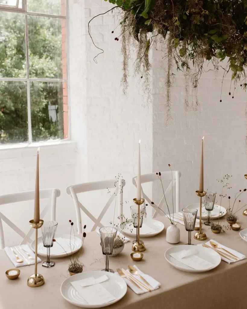 Table set for a meal with white plates, gold cutlery, glassware, beige candles, and simple floral decor beneath a hanging greenery arrangement by a large window—embrace Minimalist Magic: How to Style a Simplified Winter Table Decor.