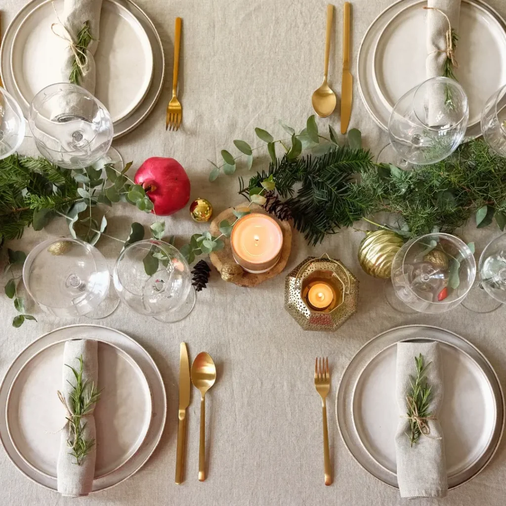 A table set for four with gold cutlery, beige napkins, wine glasses, candles, pine greenery, a pomegranate, and festive ornaments as a centerpiece captures Minimalist Magic: How to Style a Simplified Winter Table Decor.