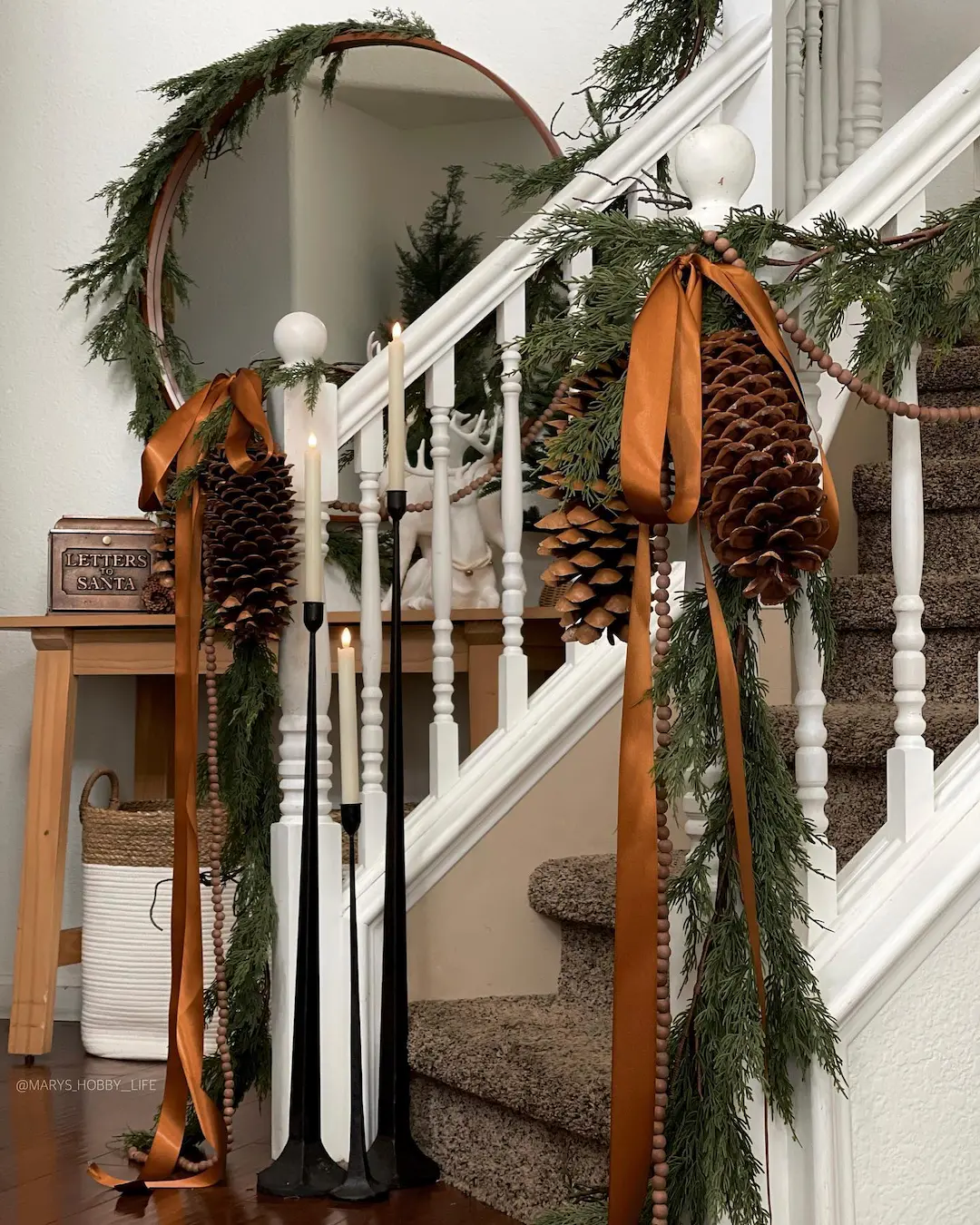 A staircase decorated with pine garland, large pinecones, and brown ribbon captures Pinecone Magic: Transforming Your Home for a Festive Christmas, with tall candles and a round mirror reflecting the enchanting greenery.