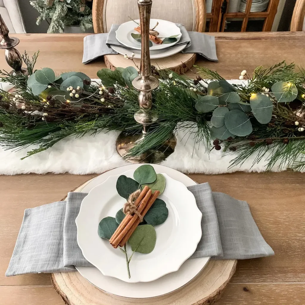 A dining table set with white plates, gray napkins, cinnamon sticks, and greenery showcases Minimalist Magic: How to Style a Simplified Winter Table Decor, featuring a pine and eucalyptus centerpiece on a wooden table.