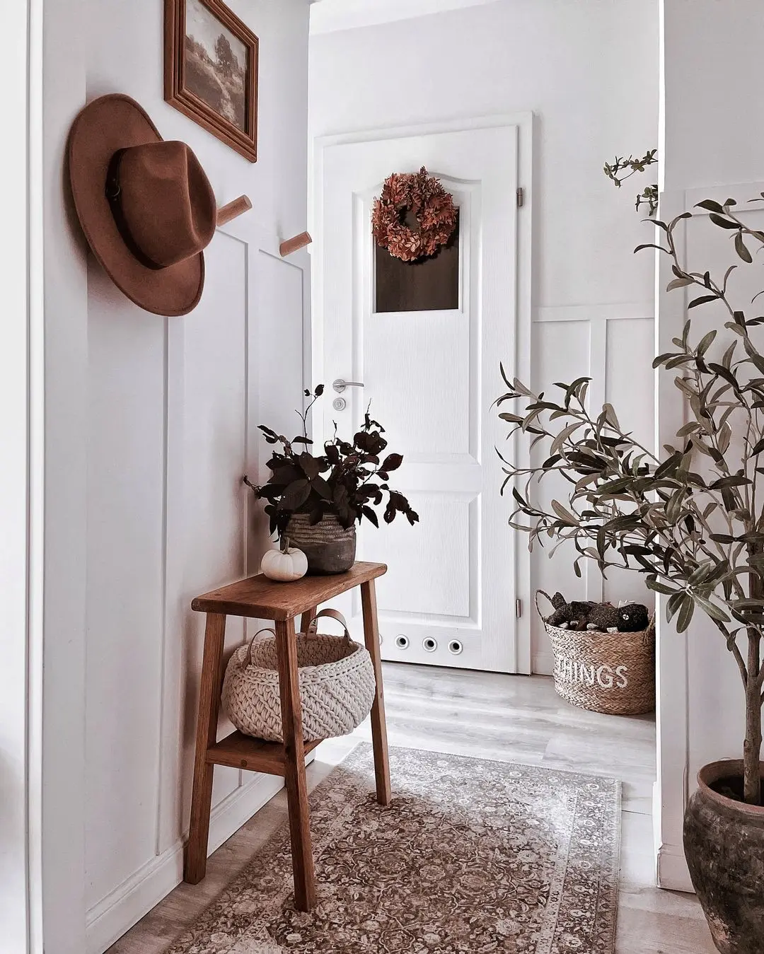 A hallway with a wooden table, hat on the wall, potted plants, decorative pillow, and basket effortlessly showcases how to avoid common mistakes when decorating for seasonal changes. A white door adorned with a wreath adds the perfect touch in the background.